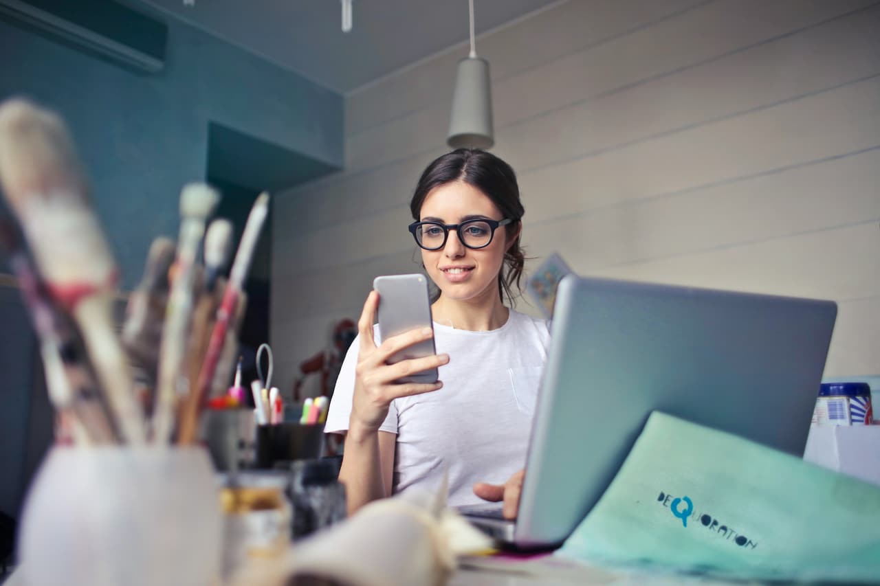 A woman at her desk reading her phone beside her laptop