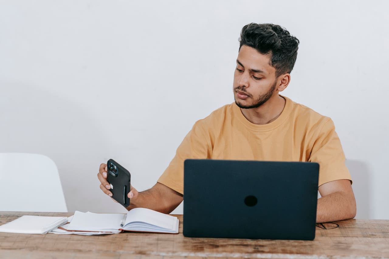A Latino man at his laptop checking a message on his phone